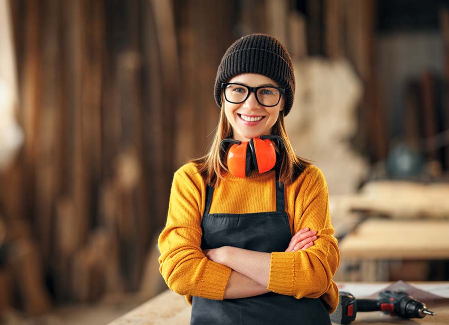 Smiling confident young female joiner in apron standing near workbench and looking at camera friendly while working in craft workshop